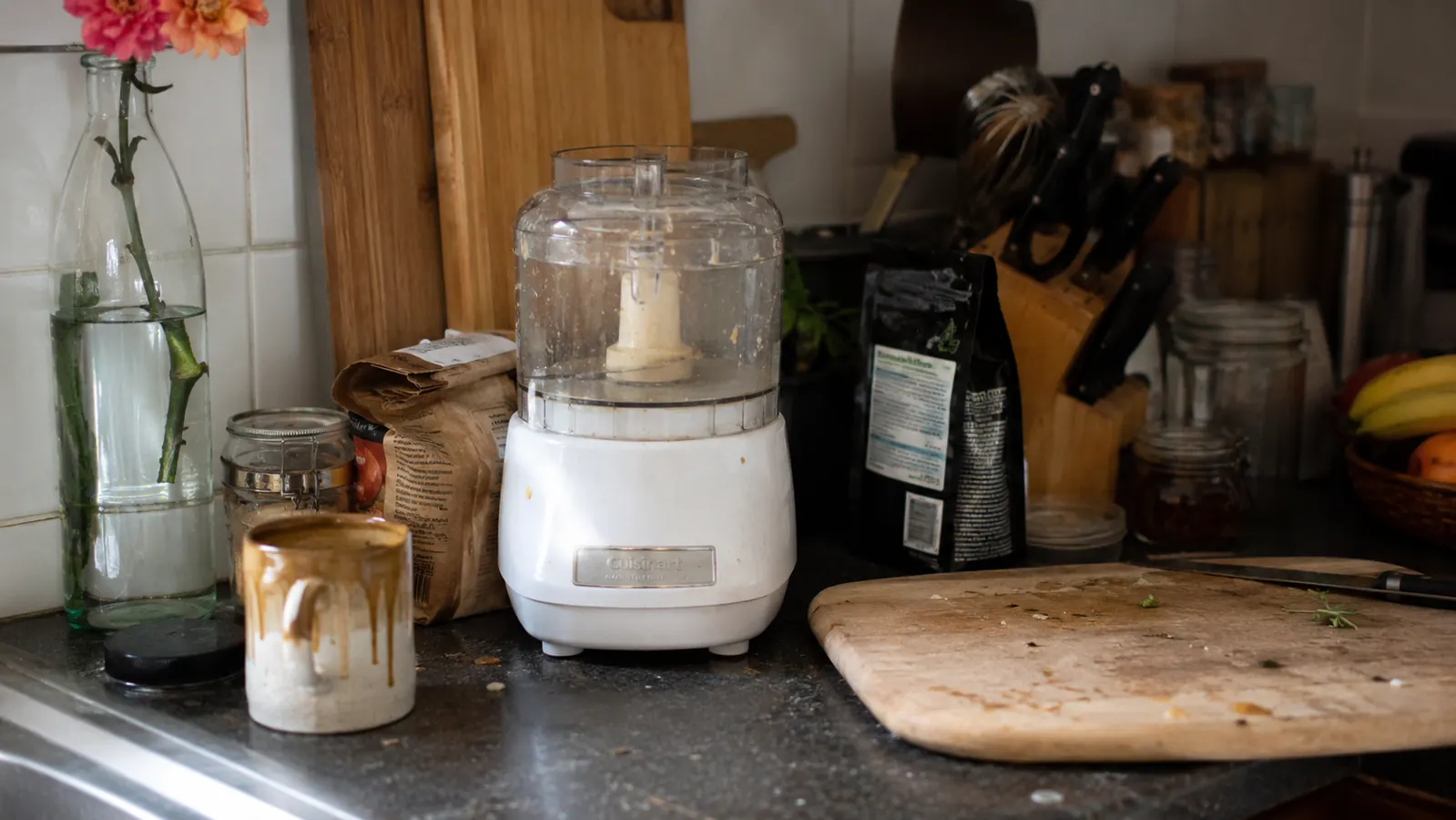 Small kitchen counter at dawn with a compact white food processor sitting next to a coffee mug and a cutting board