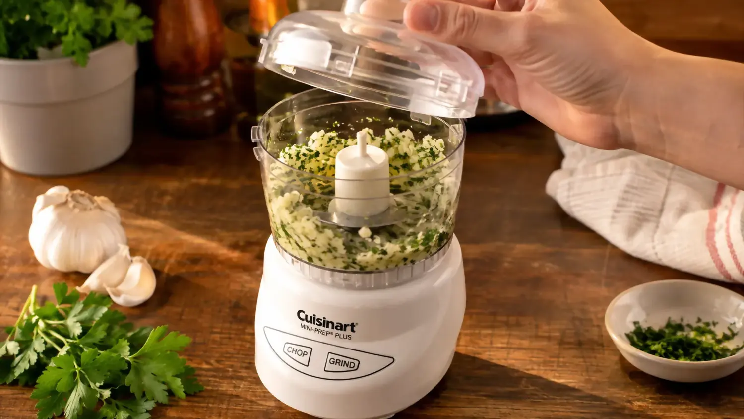 Hand pulling the lid off a small white food processor after mincing garlic and parsley inside