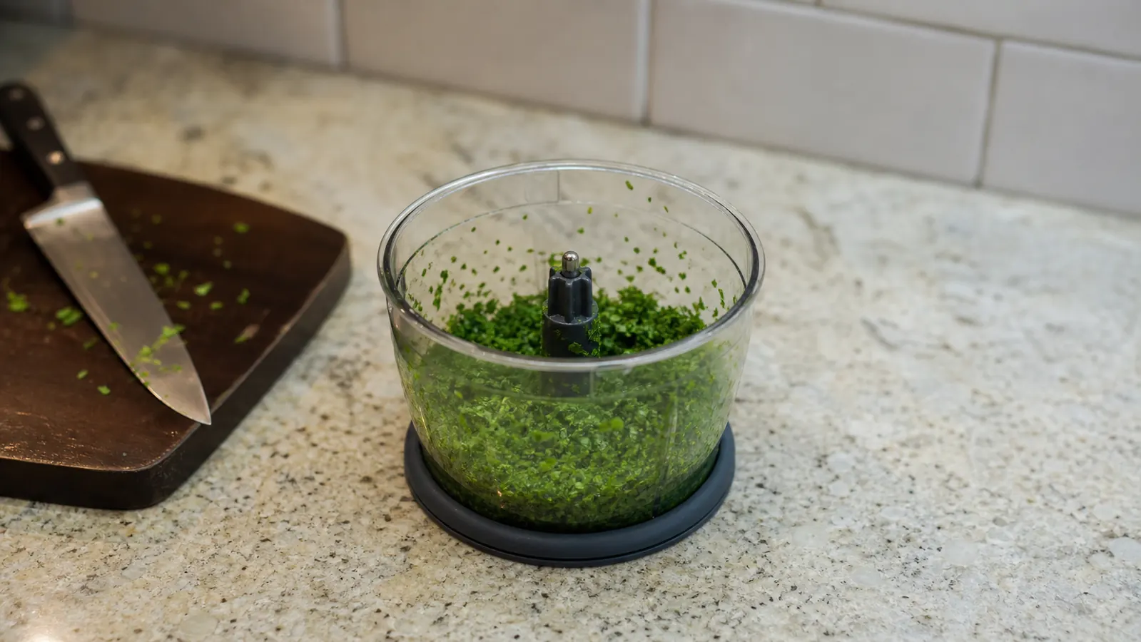 A small food processor bowl full of finely chopped parsley sitting on a compact kitchen counter next to a cutting board