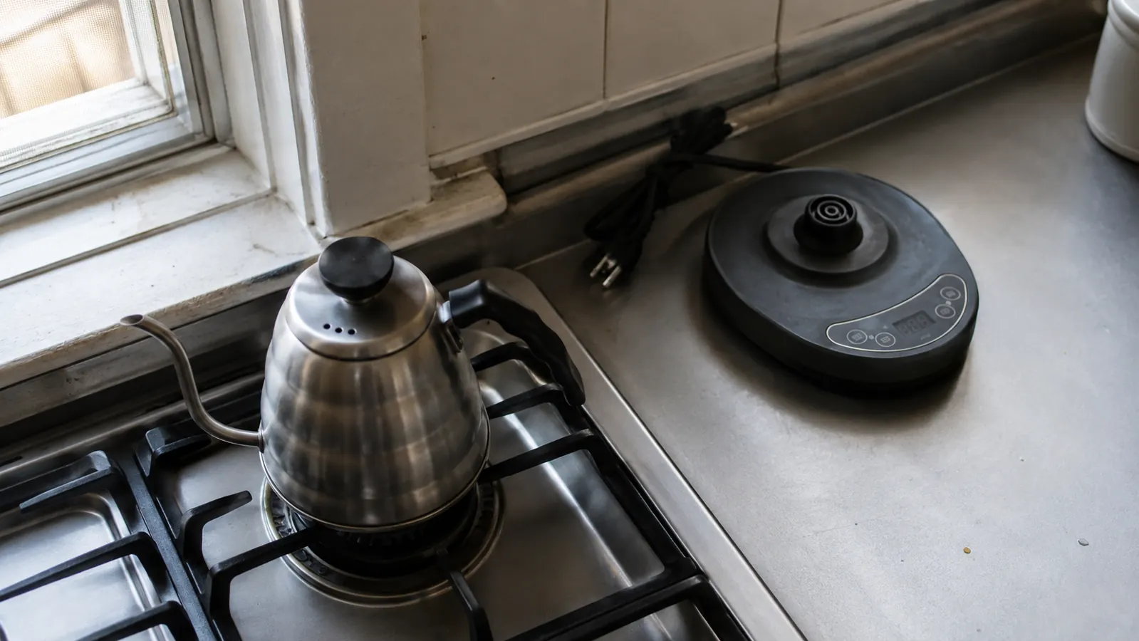 A plain stovetop gooseneck kettle sitting on a stove burner next to an unplugged electric kettle base